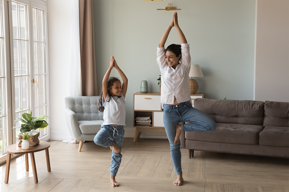A parent and child do a tree pose while exercising in their living room.