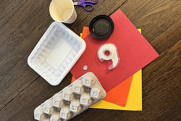 Pile of craft supplies including egg carton, tape, scissors, colored paper, cup and lid, etc.